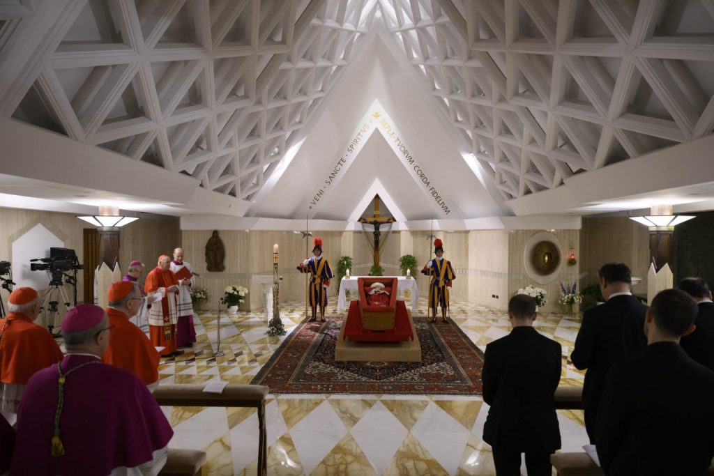 El papa descansa sobre un féretro de madera forrado de terciopelo rojo con una casulla purpura y una mitra blanca y en las manos lleva un rosario