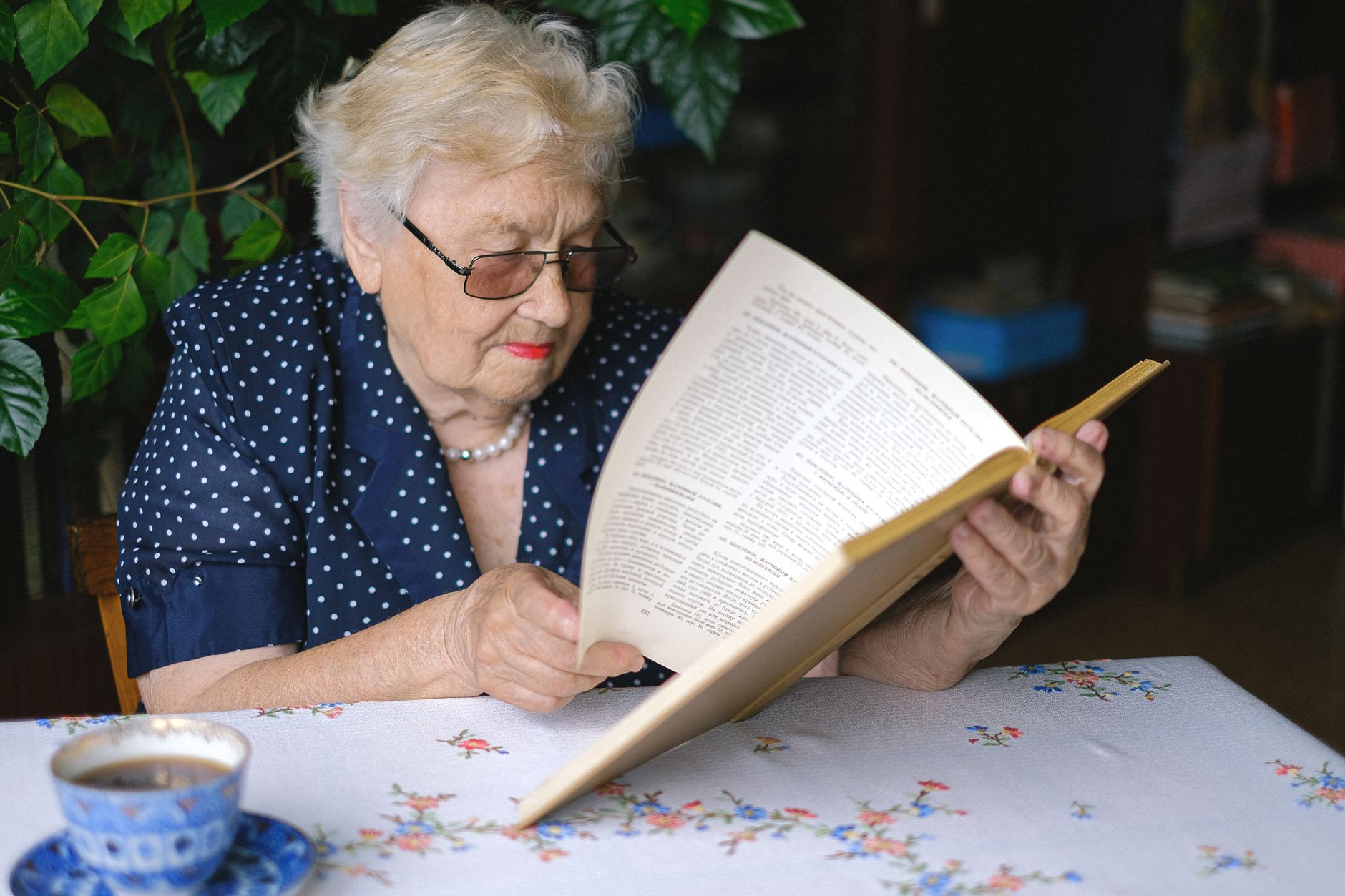 Mulher idosa de óculos lendo um livro em casa, desfrutando do tempo de lazer com chá.