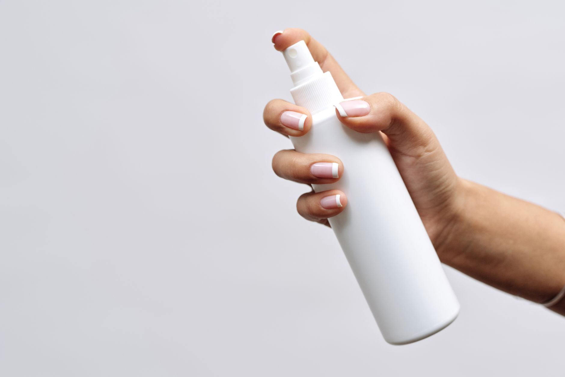 Uma mão com unhas bem cuidadas segura uma garrafa spray branca minimalista em um fundo branco, perfeita para mockups de branding.