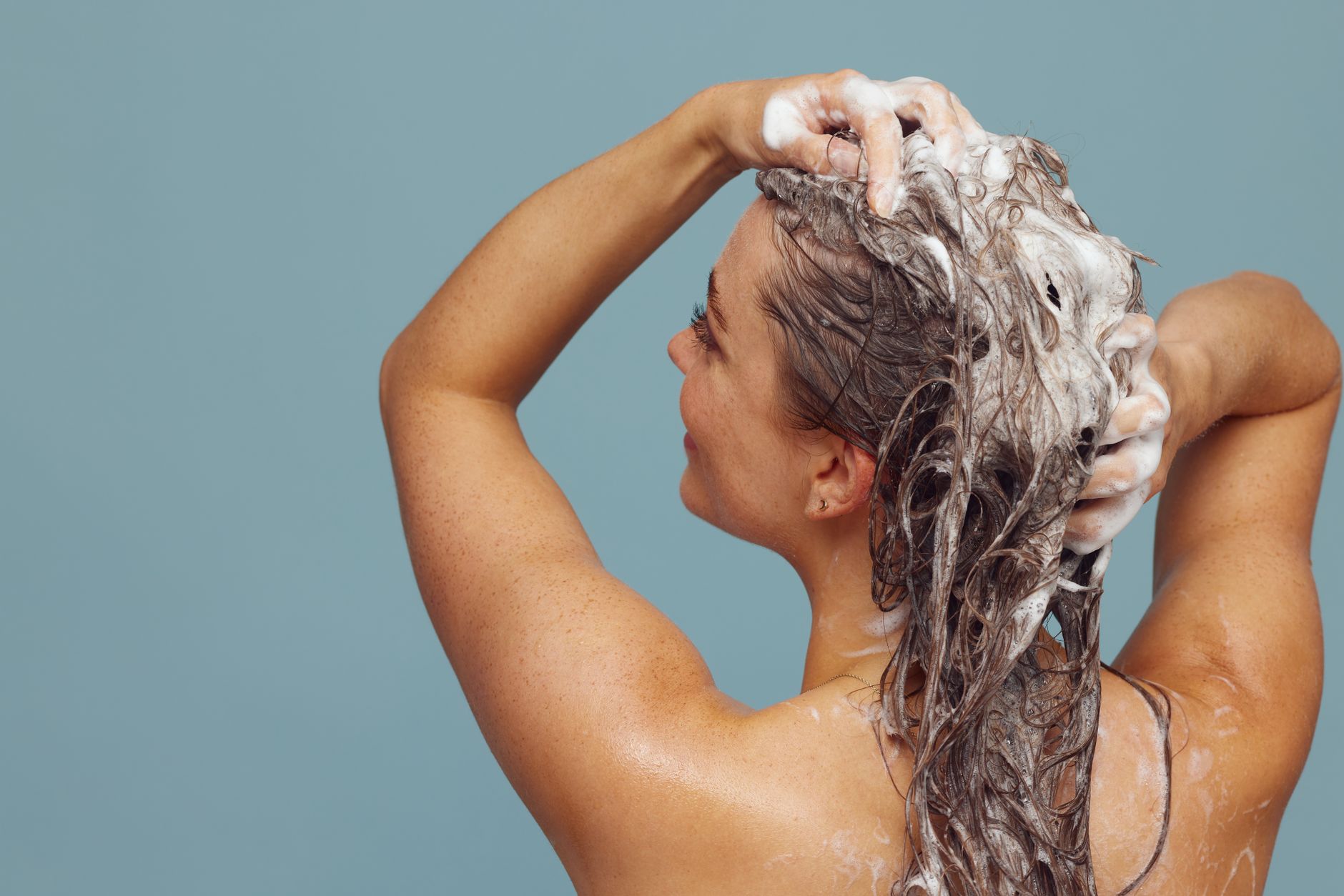 Mulher com cabelo longo lavando-o com shampoo, criando espuma, em ambiente interno contra fundo azul.