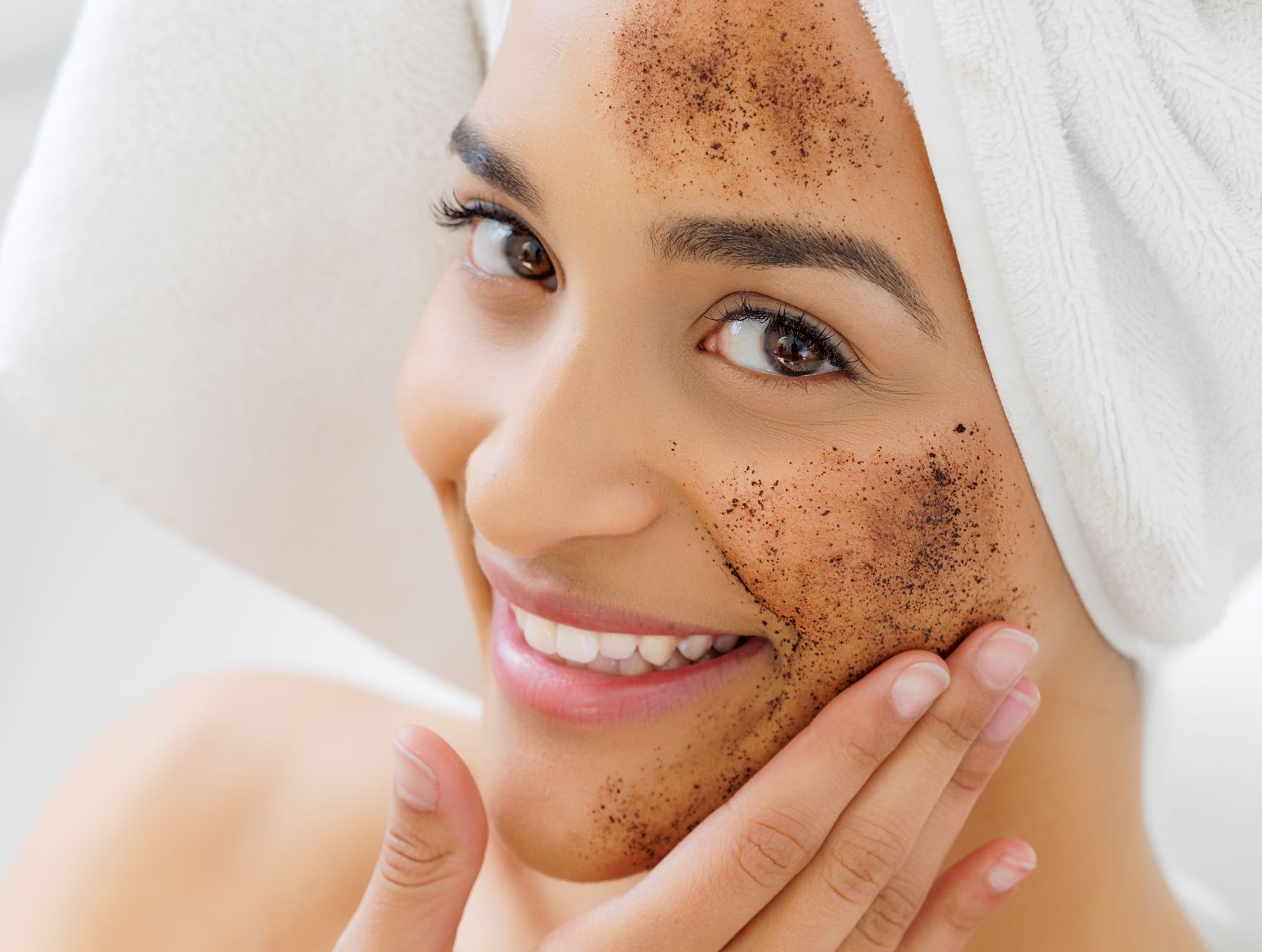 Mulher sorridente com toalha na cabeça aplicando uma máscara facial esfoliante natural. Conceito de cuidados com a pele e autocuidado.