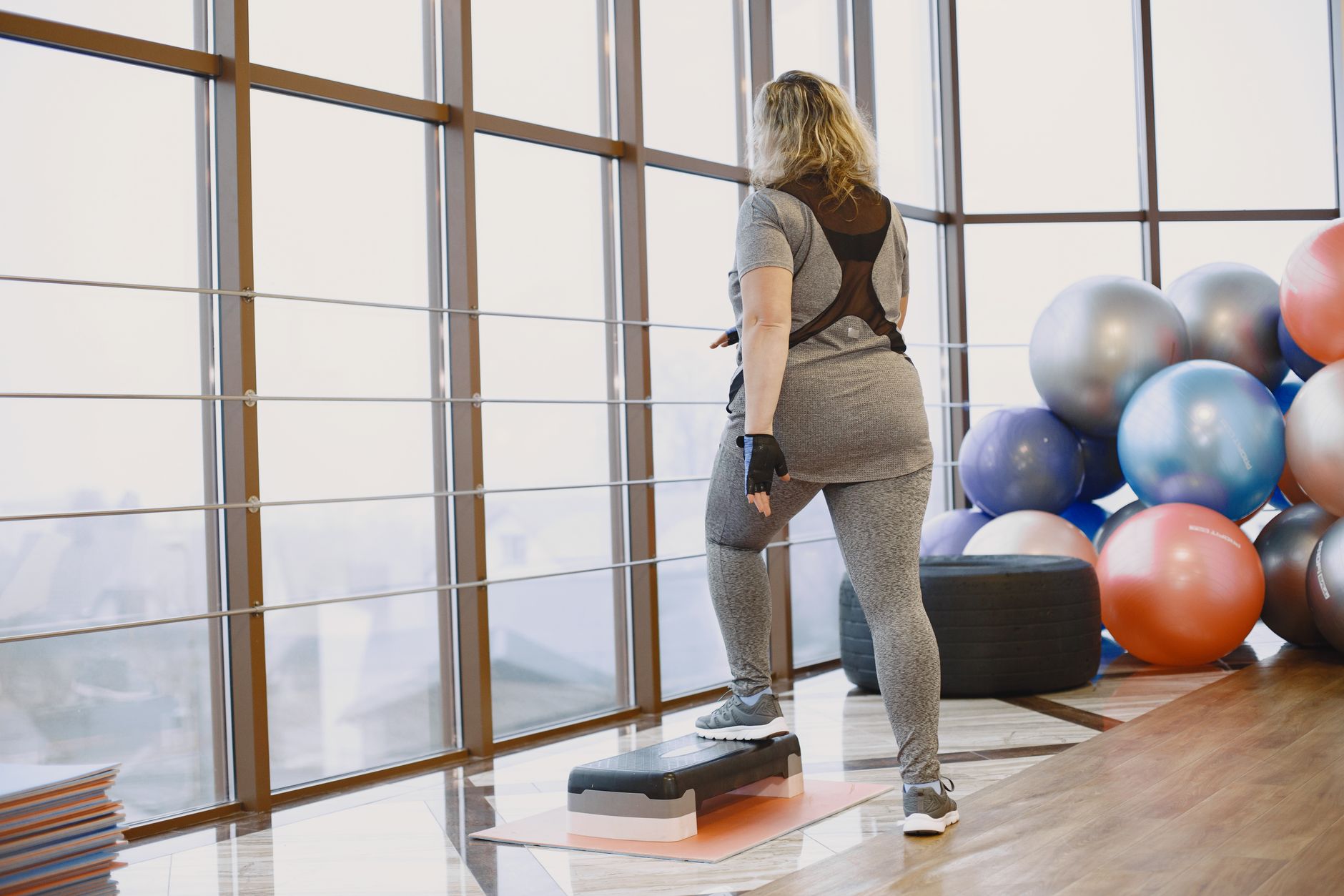 Vista traseira de uma mulher se exercitando em ambientes fechados com equipamentos de fitness e plataforma de step.