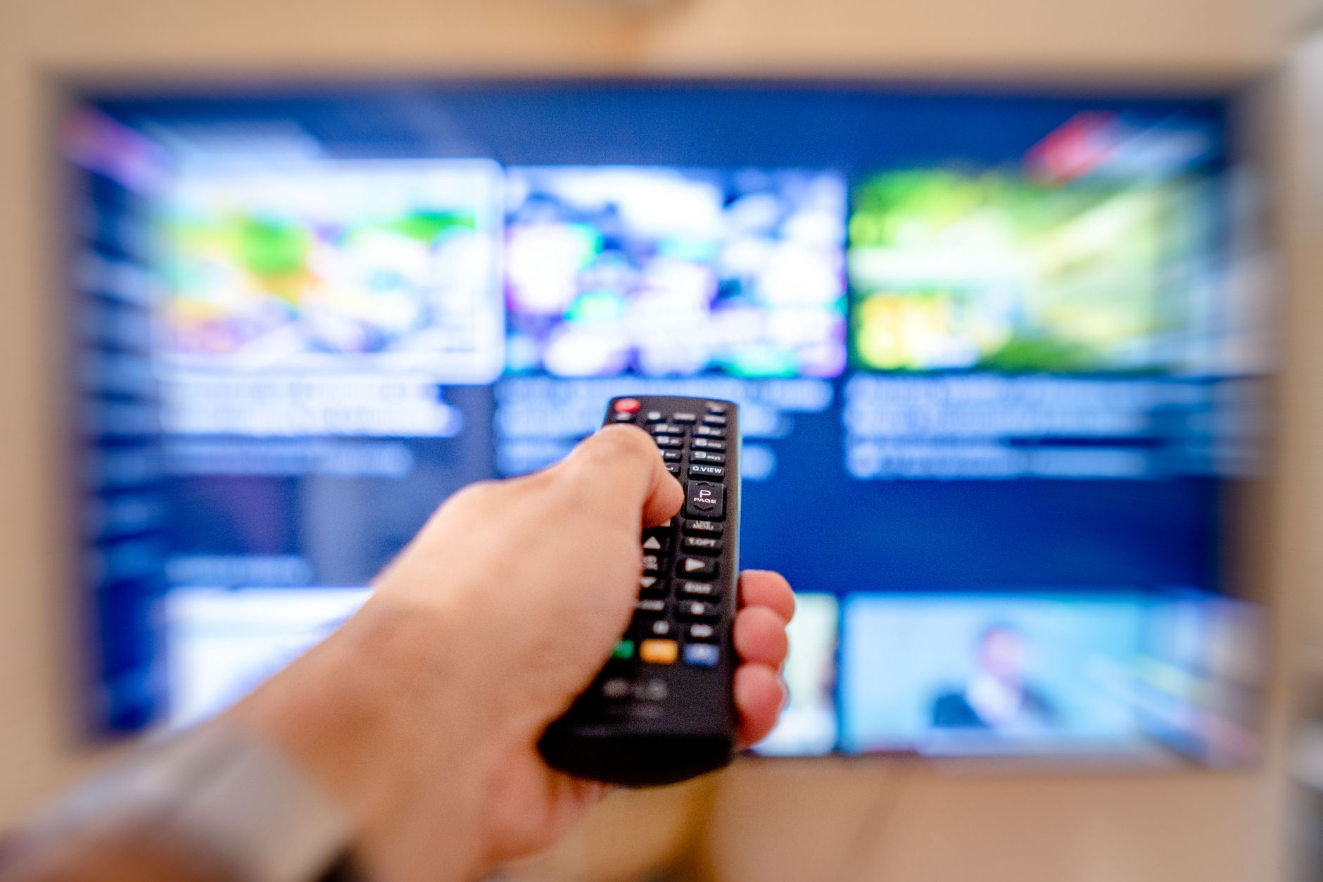 Mão segurando o controle remoto apontando para uma tela de TV desfocada em um ambiente interno aconchegante.