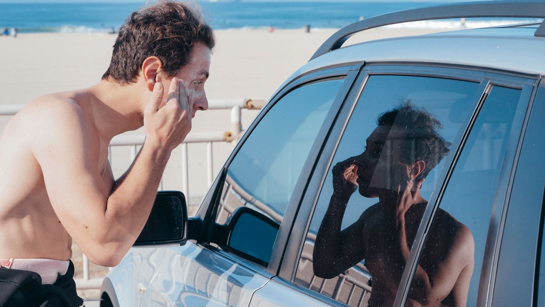 Um homem aplica protetor solar usando o reflexo da janela do carro em uma praia ensolarada.