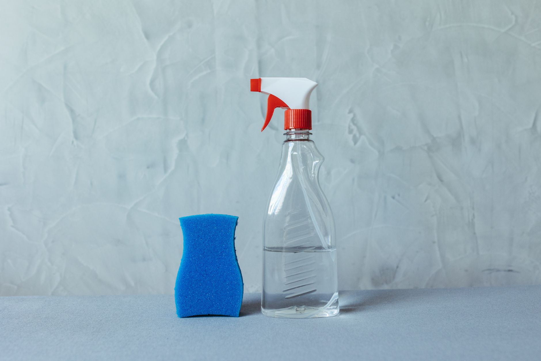 Um frasco spray e uma esponja azul sobre uma superfície cinza texturizada. Perfeito para temas de limpeza.